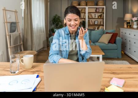 Freelance businesswoman in home office reviewing charts and dictating a voice note while analyzing financial reports on a laptop — remote work, strate Stock Photo
