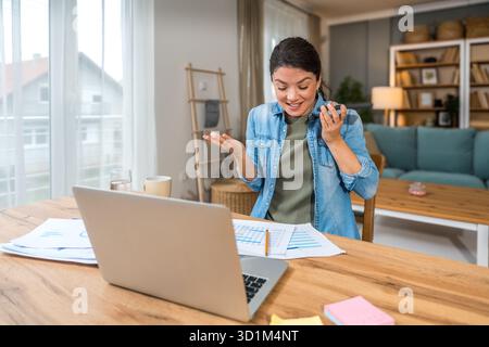 Freelance businesswoman in home office reviewing charts and dictating a voice note while analyzing financial reports on a laptop — remote work, strate Stock Photo