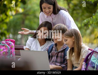 Schoolchildren happily looking at laptop while sitting in nature on bench while teacher explaining something behind them Stock Photo