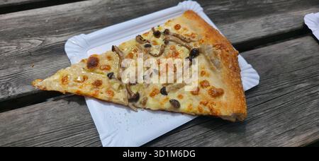 Gourmet pizza slice topped with mushrooms, placed on a white paper plate, resting on a rustic wooden table, showcasing appetizing textures and colors. Stock Photo
