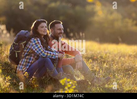 Two hikers sit together on grassy patch, smiling and sharing a peaceful moment as the sun sets, surrounded by the beauty of nature in autumn Stock Photo