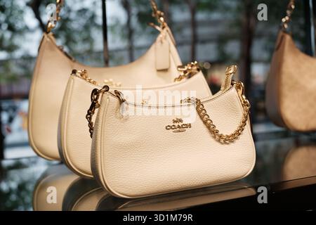 Coach branded leather handbags in white are arranged on display, featuring gold chain details and a shiny logo against a blurred outdoor backdrop. Stock Photo