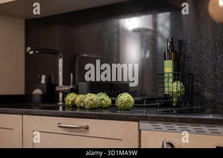 A modern kitchen countertop with a black granite surface. Fresh green artichokes are arranged beside a bottle of olive oil in a wire basket. Stock Photo
