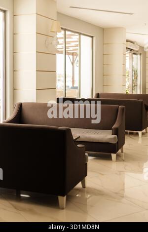 Cozy lounge area features several brown sofas arranged in an inviting manner. Natural light floods in through large windows, creating a warm atmospher Stock Photo