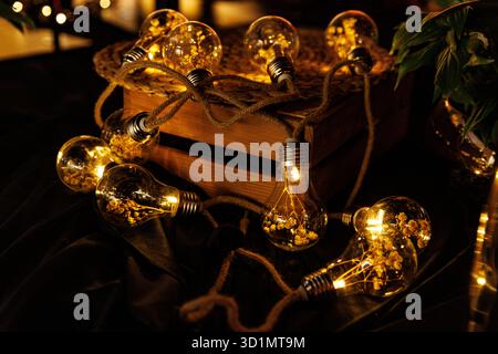 String of decorative light bulbs with glowing filaments placed on a wooden box and dark fabric, creating warm ambient light. Stock Photo