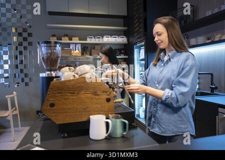 Serious concentrated Caucasian barista operating the coffee machine next to her pleased Asian female colleague Stock Photo