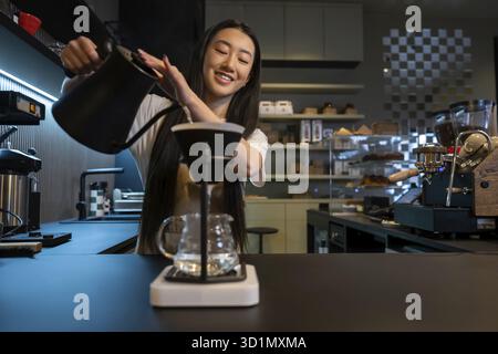 Smiling barista pouring hot water from the kettle through the filter inside the pour-over coffee maker funnel Stock Photo