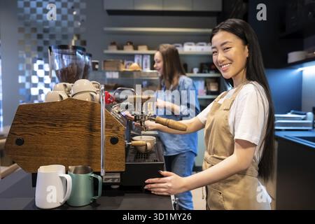 Smiling barista making coffee with the espresso machine in a cafe in the presence of her female colleague Stock Photo