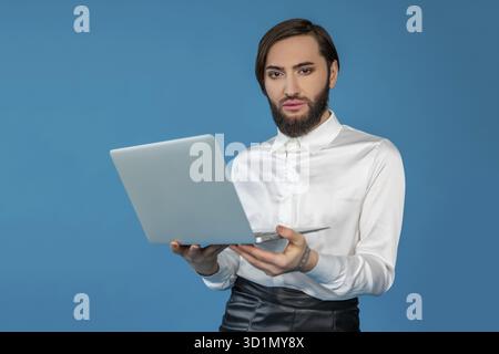 Self confident transgender man wearing white shirt working on laptop browsing internet looking at camera posing isolated over blue background Stock Photo