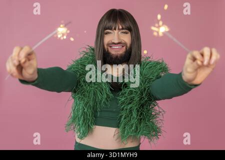 Joyful transsexual with makeup and wig wearing green feather dress having fun with sparklers, spending leisure time on party posing isolated over pink Stock Photo