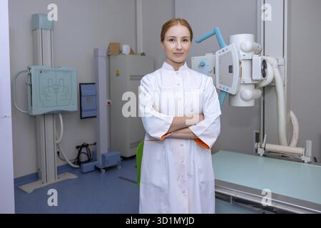 Caucasian blonde female doctor radiologist wearing lab coat posing near X-ray machine in hospital Stock Photo