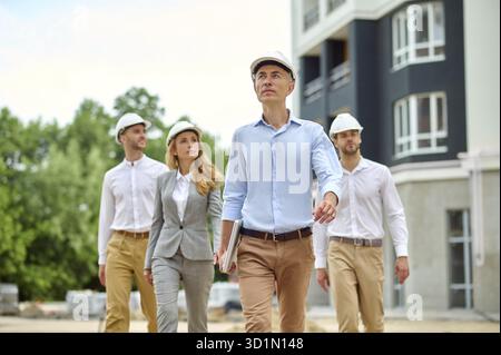 Calm Caucasian male engineer followed by a female construction inspector and two builders in hardhats Stock Photo