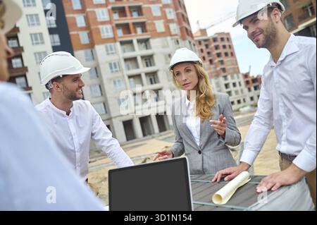Serious middle-aged female building inspector standing on the construction site surrounded by three male workers Stock Photo