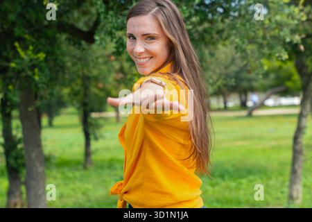 Smiling woman in yellow shirt taking selfie with red smartphone, lying on grass. Stock Photo