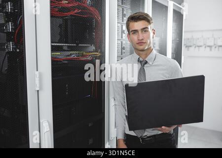 Portrait of modern young man holding laptop standing in server room working with supercomputer Stock Photo