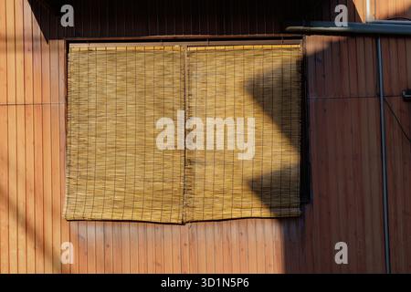 Sunlight casts pronounced shadow on bamboo blinds that cover a window on wood pattern building, creating abstract image resembling an open mouth. Stock Photo