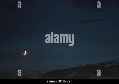 Partly obscured by cloud's thin veil, crescent moon shines with Venus on deep blue backdrop of cloudy evening sky, creating peaceful lunar atmosphere. Stock Photo
