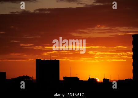 Dawn sun sliver appears over silhouetted building, illuminating sky and lingering clouds in orange and blood red hues ,creating captivating cityscape. Stock Photo