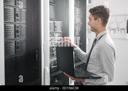 Male Server Engineer Works on a Laptop in Large Data Center Stock Photo