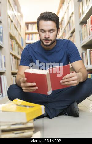 Handsome young student sitting on library floor reading book in college Stock Photo