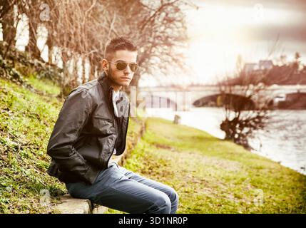 Three-quarter length of contemplative light brown haired young man wearing grey jacket and denim jeans sitting on wall beside picturesque river in Tur Stock Photo