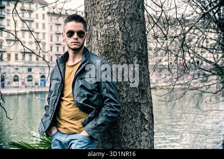 Three-quarter length of contemplative light brown haired young man wearing grey jacket and denim jeans standing beside picturesque river in Turin, Ita Stock Photo