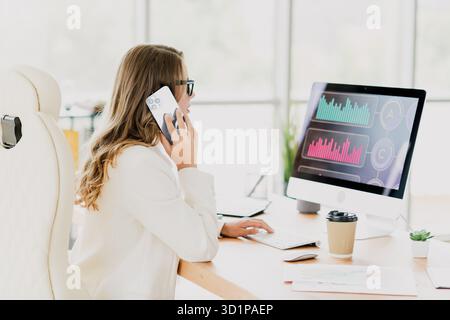 Businesswoman discussing analytics on smartphone in modern office with vibrant graphs displayed on computer screen Stock Photo