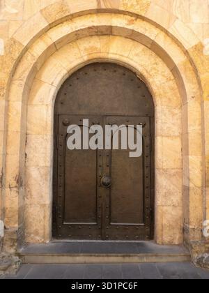 Old arched wooden door in a stone Orthodox church facade, with warm sunlight and historic texture in Tbilisi. Stock Photo