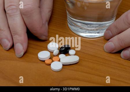 Hands are placed near assorted pills and a glass of water on a wooden surface. Stock Photo