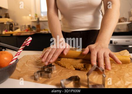 A person is using a wooden rolling pin to flatten gingerbread cookie dough on parchment paper in the kitchen. Cookie cutters are visible and a candy cane and orange are nearby. Stock Photo