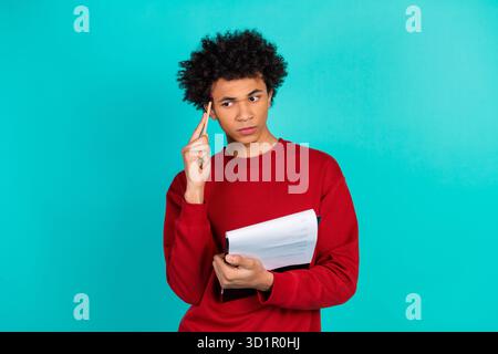 Young man in red sweater holding papers and pencil stands against teal background looking thoughtful and stylish Stock Photo