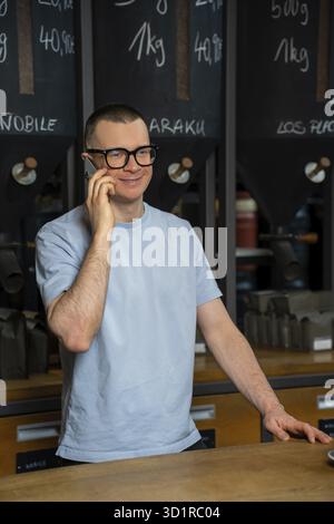 Handsome barista in glasses talking on mobile phone in coffee shop, bartender coffeemaker at his workplace Stock Photo
