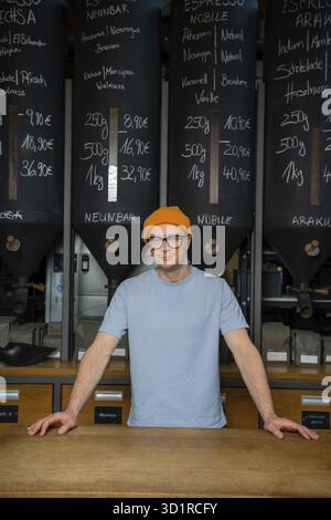 Handsome hipster bartender wearing beanie hat and glasses in the interior of modern coffee shop with big variety of coffee beans Stock Photo