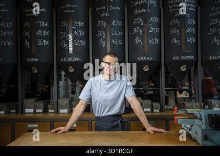 Smiling young entrepreneur wearing apron and glasses standing near counter of his cafe with big variety of coffee Stock Photo