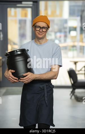 Young hipster barista wearing hat, glasses and apron carrying barrel preparing for work in coffee shop Stock Photo