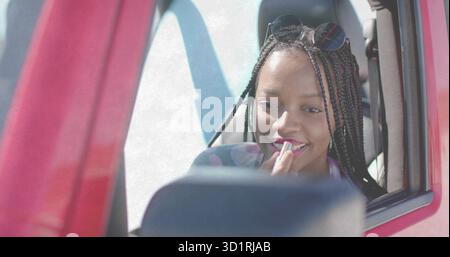 Woman wearing sunglasses applying lipstick from tube and looking into side mirror, in red car Stock Photo