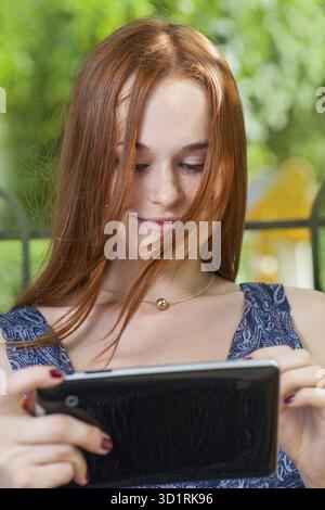 Redhead student leaning against a tree using her tablet on college campus. Redhead woman with tablet outdoors Stock Photo