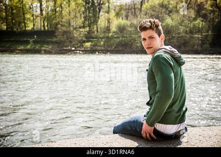 Three-quarter length of contemplative light brown haired young man wearing green hooded-shirt and denim jeans sitting on wall beside picturesque river Stock Photo