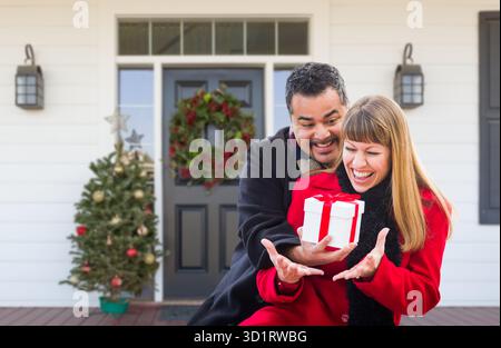 Young Mixed Race Couple Exchanging Gift On Front Porch of House with Christmas Decorations Stock Photo