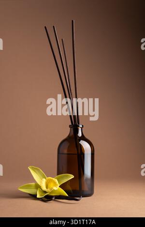 Reed diffuser with vanilla sticks in a brown glass bottle and vanilla flower on brown background. Minimal composition symbolizing calmness, purity, an Stock Photo