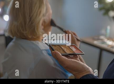 In a cozy beauty salon, the artist delicately applies cream to a middle-aged blonde woman’s face with a brush, delivering an even and professional res Stock Photo