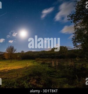 Vast night sky filled with stars and a bright full moon, with a clear satellite trail passing over a rural field and trees Stock Photo