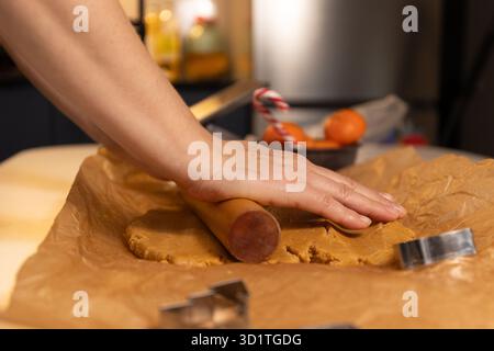 In a kitchen, hands are rolling out gingerbread dough on parchment paper with a wooden rolling pin. Cookie cutters are visible, ready for cutting shapes. Celebrating Christmas with baking. Stock Photo