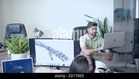 Working Indian man typing on laptop at open office desk, with robotic hand schematic, copy space Stock Photo