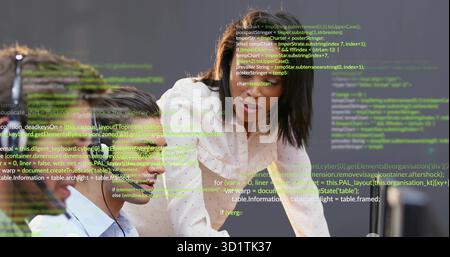 Leaning Black woman guiding seated colleague at office desk with headphones, floating code overlay Stock Photo