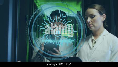 Two technicians in lab coats analyzing tablet in data center aisle, with holographic interface Stock Photo