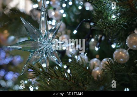 Transparent acrylic star ornament hangs on a Christmas tree. The star reflects light from nearby fairy lights. Surrounding the star are evergreen bran Stock Photo