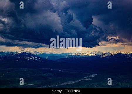 Ominous storm clouds hang over the Grand Teton mountain range in Wyoming, with snow-capped peaks and a winding river valley below. Stock Photo