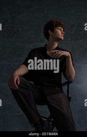 young man with curly hair sits confidently in a black shirt leaning slightly on a chair. The studio backdrop adds depth emphasizing his relaxed pose and contemplative mood. Stock Photo