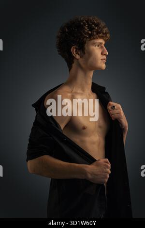 young man with curly hair stands in profile in a studio setting. He wears a black shirt slightly pulled back showcasing his relaxed pose. The lighting creates depth and highlights his features. Stock Photo